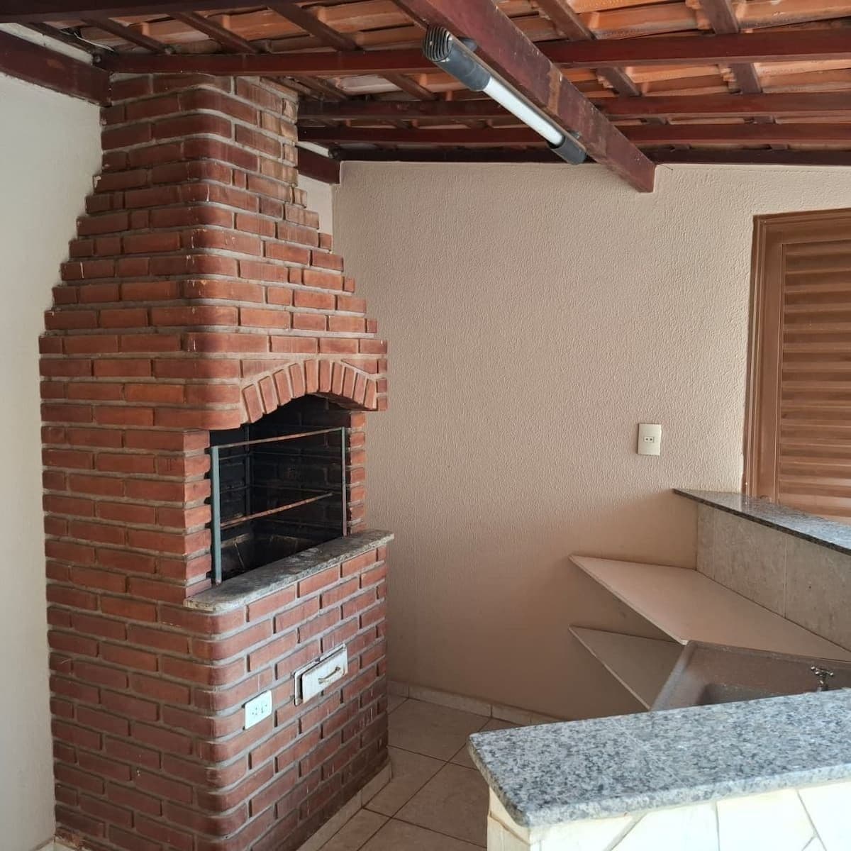 A brick-built outdoor fireplace with a stone hearth, a wooden ceiling, and a tiled floor.