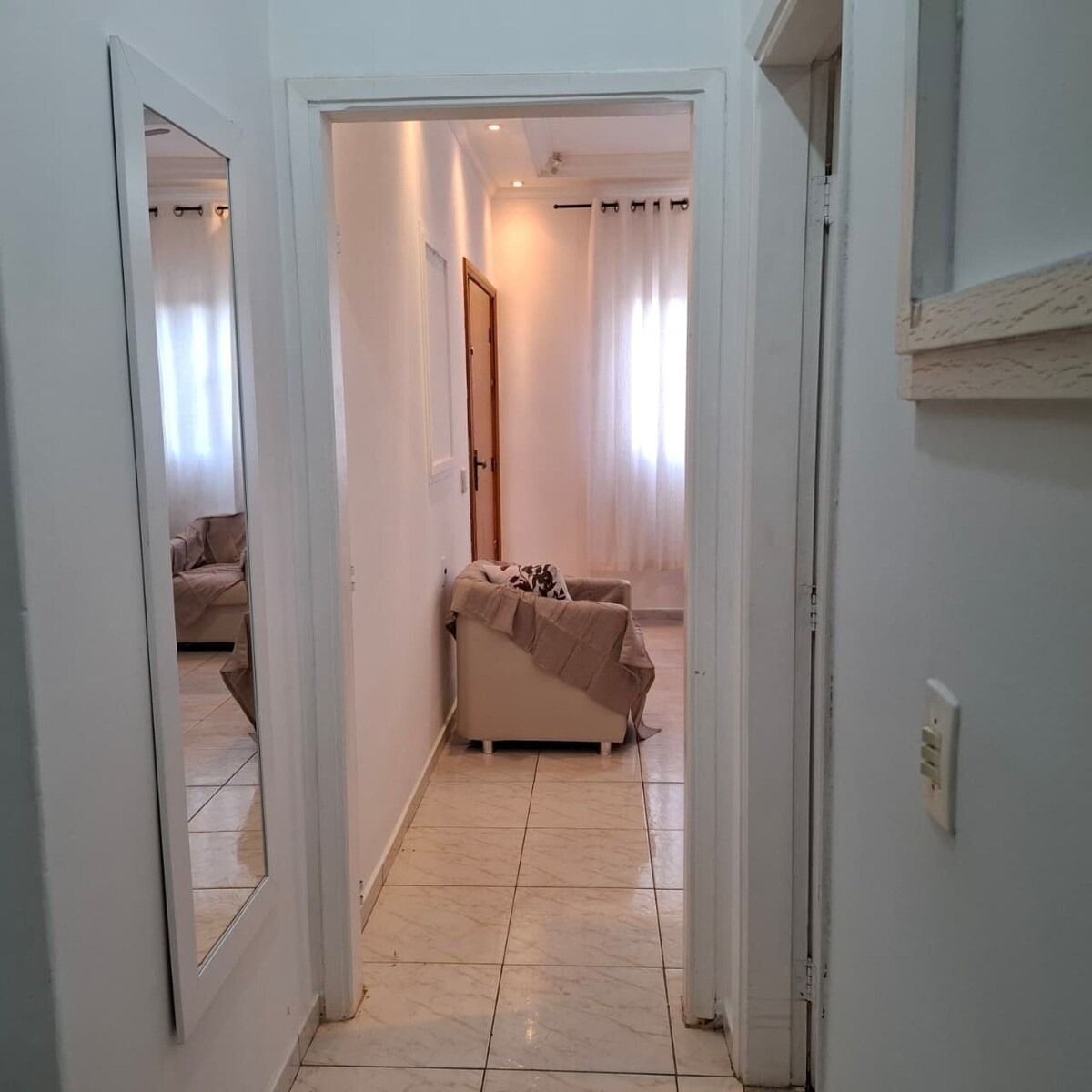 A hallway with a bed, a door, and a mirror reflecting a room with a bed.