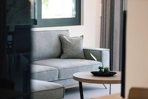 Un salon moderne avec un canapé gris, une petite table ronde ornée d’une plante et une fenêtre avec des rideaux.