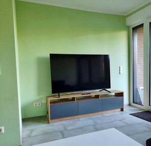 Un saló modern amb un televisor de pantalla plana sobre un moble de fusta, parets de verd clar i una porta corredissa de vidre que dona a una zona exterior.