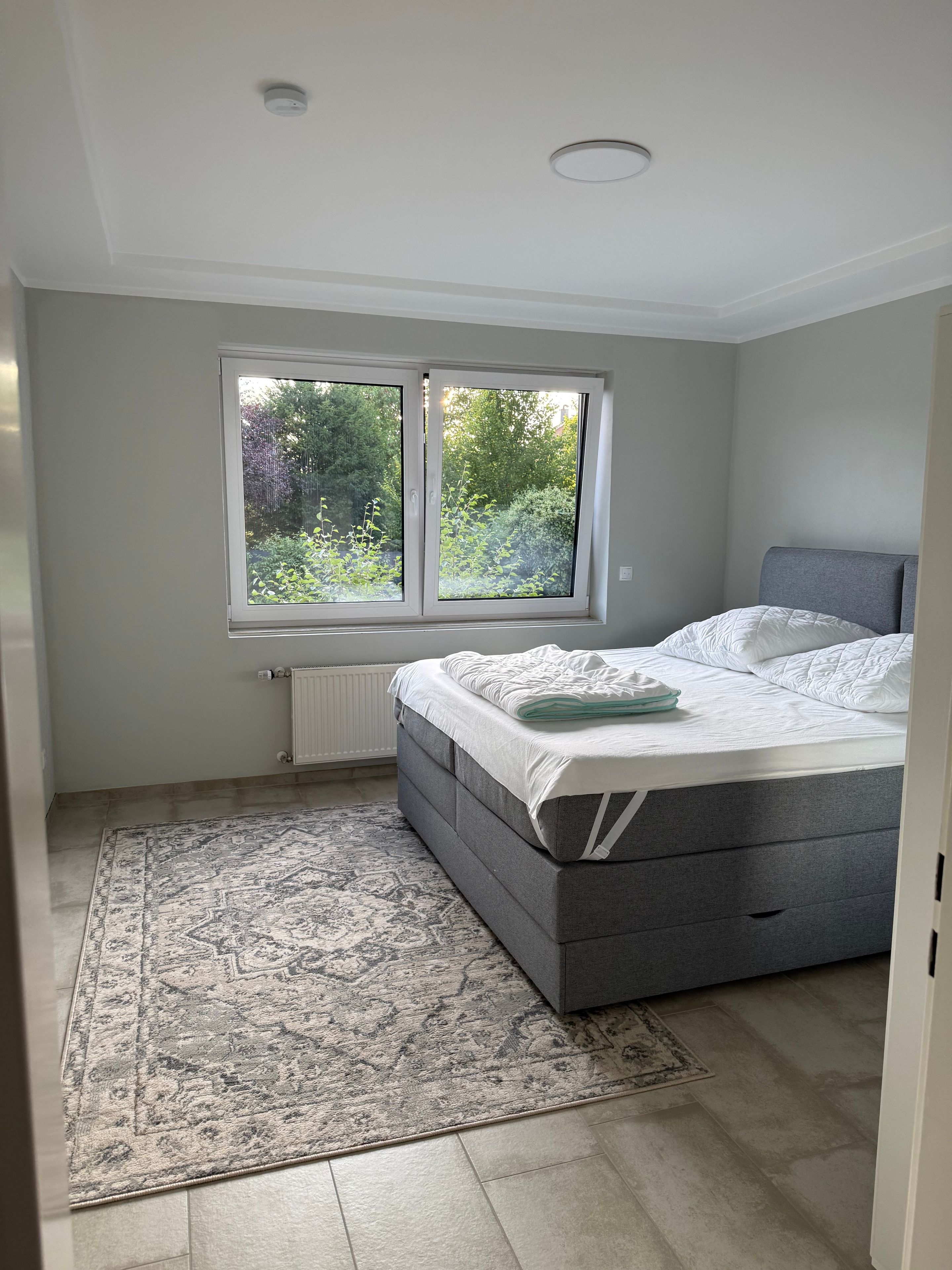 Ein Schlafzimmer mit einem Bett, einem Fenster, einer Heizung und einem gemusterten Teppich.