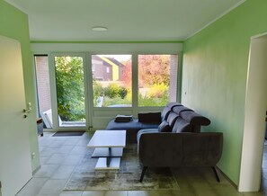 Habitació amb un sofà, una taula i vista a un jardí a través d'una porta de vidre.