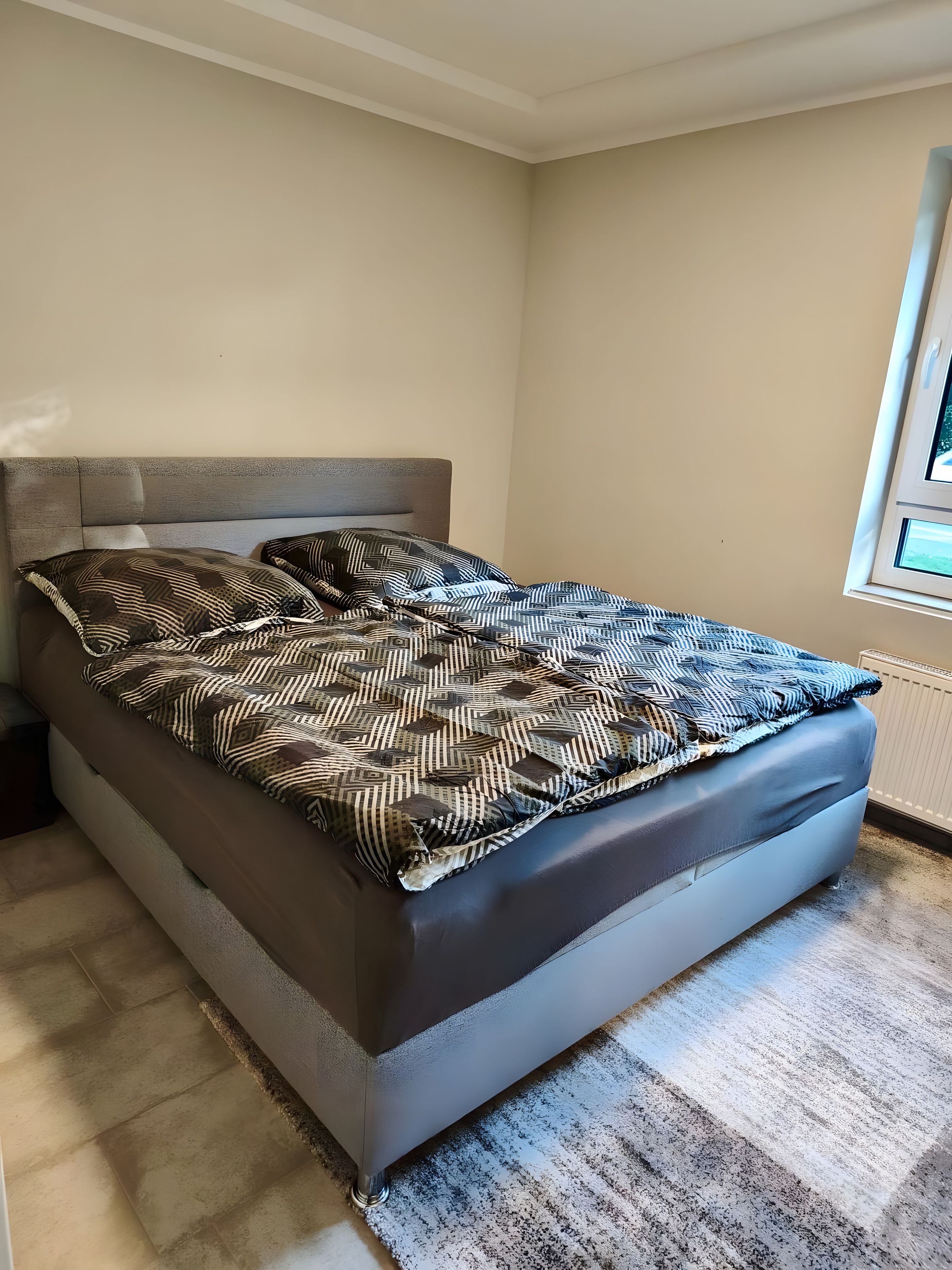 Een bed met een beddegoed met patroon en kussens, een hoofdbord en een raam met uitzicht.