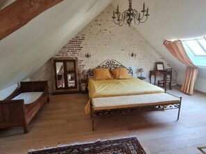 Een slaapkamer met een schuine kap, een bed met hoofdeinde, een houten bankje, een kleine tafel, een stoel, een kroonluchter en een raam met gordijnen.
