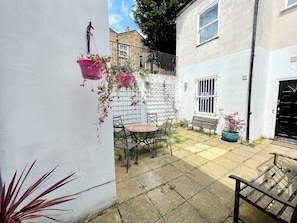 A courtyard with a white wall, a metal table with chairs, a bench, and hanging flower pots. - Bright Home Near Central London - Near Emirates Stadium (London)
