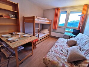 Ein Schlafraum mit Etagenbetten, einem Schreibtisch mit Geschirr und Besteck, einem Bett mit Blumendekoration auf der Bettdecke und einem Fenster mit Vorhängen.