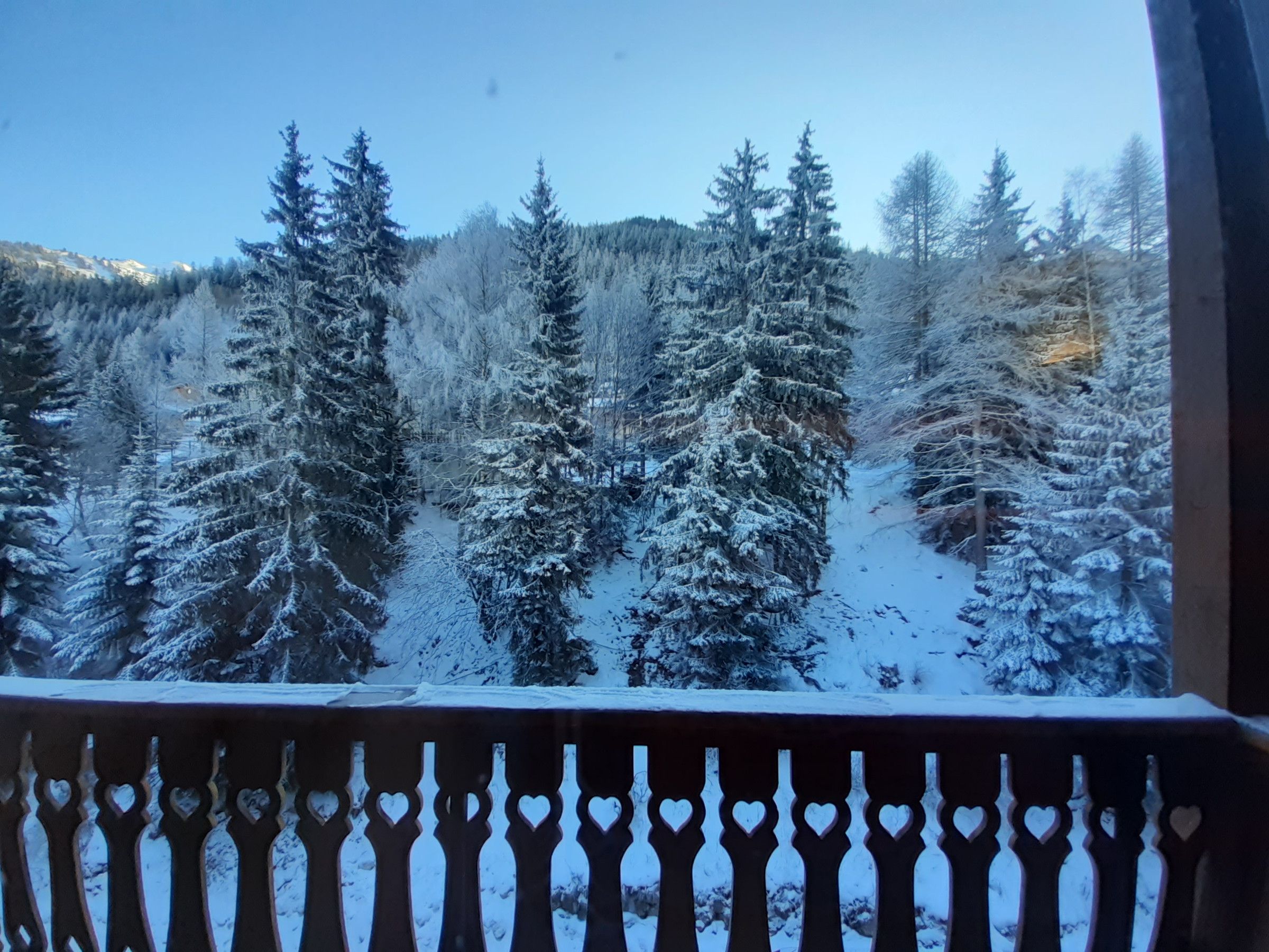 A balcony with a railing featuring heart-shaped cutouts, overlooking a snowy forest with tall evergreen trees.