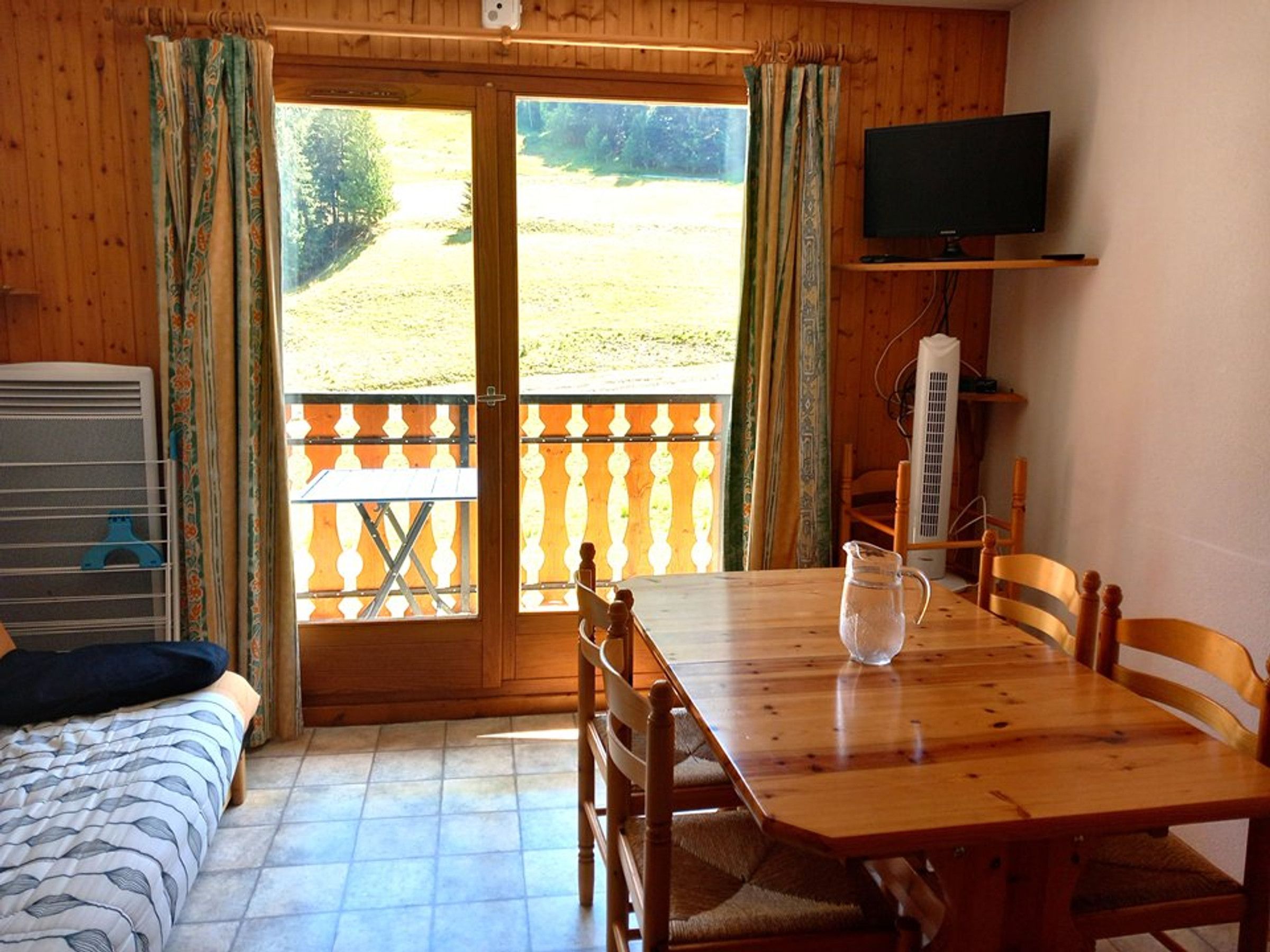 A room with a bed, a table, chairs, a TV, and a balcony with a view of the outdoors.