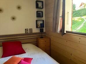 Ein Schlafzimmer mit einem Bett, einem Nachttisch, einem Fenster mit Vorhängen und an der Wand befestigten Bilderrahmen.