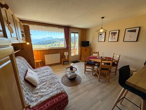 Ein gemütliches Zimmer mit einem Bett, einem Esstisch, Stühlen, einem Fernseher und einem Fenster mit Bergblick.