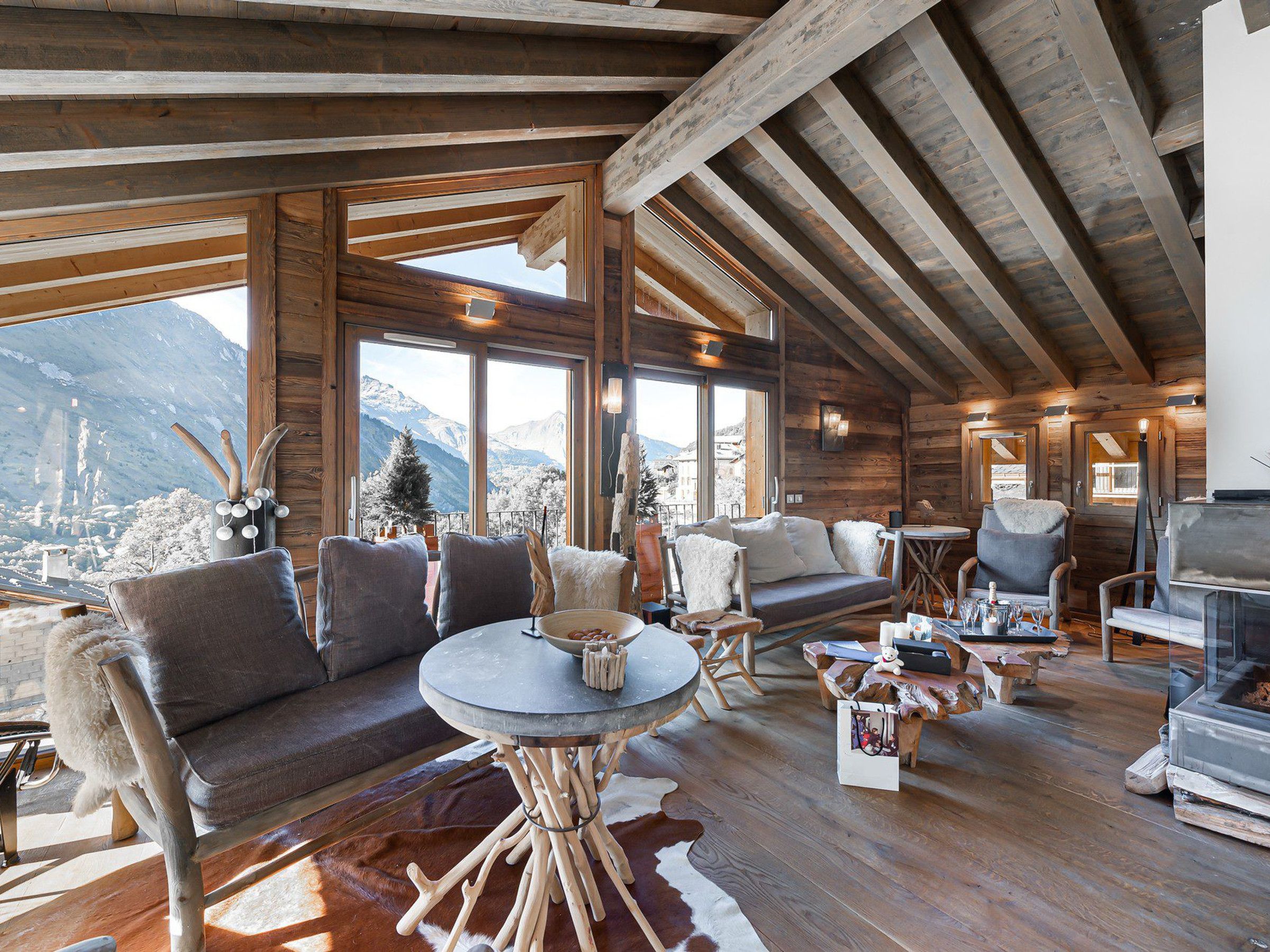A cozy cabin interior with a wooden ceiling, large windows offering mountain views, a fireplace, and rustic furniture.