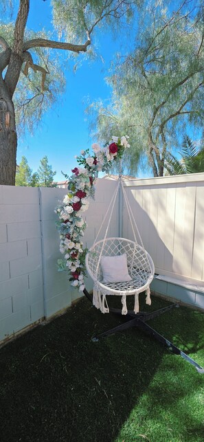 Une chaise suspendue avec un coussin, une composition florale et un arbre dans un jardin.