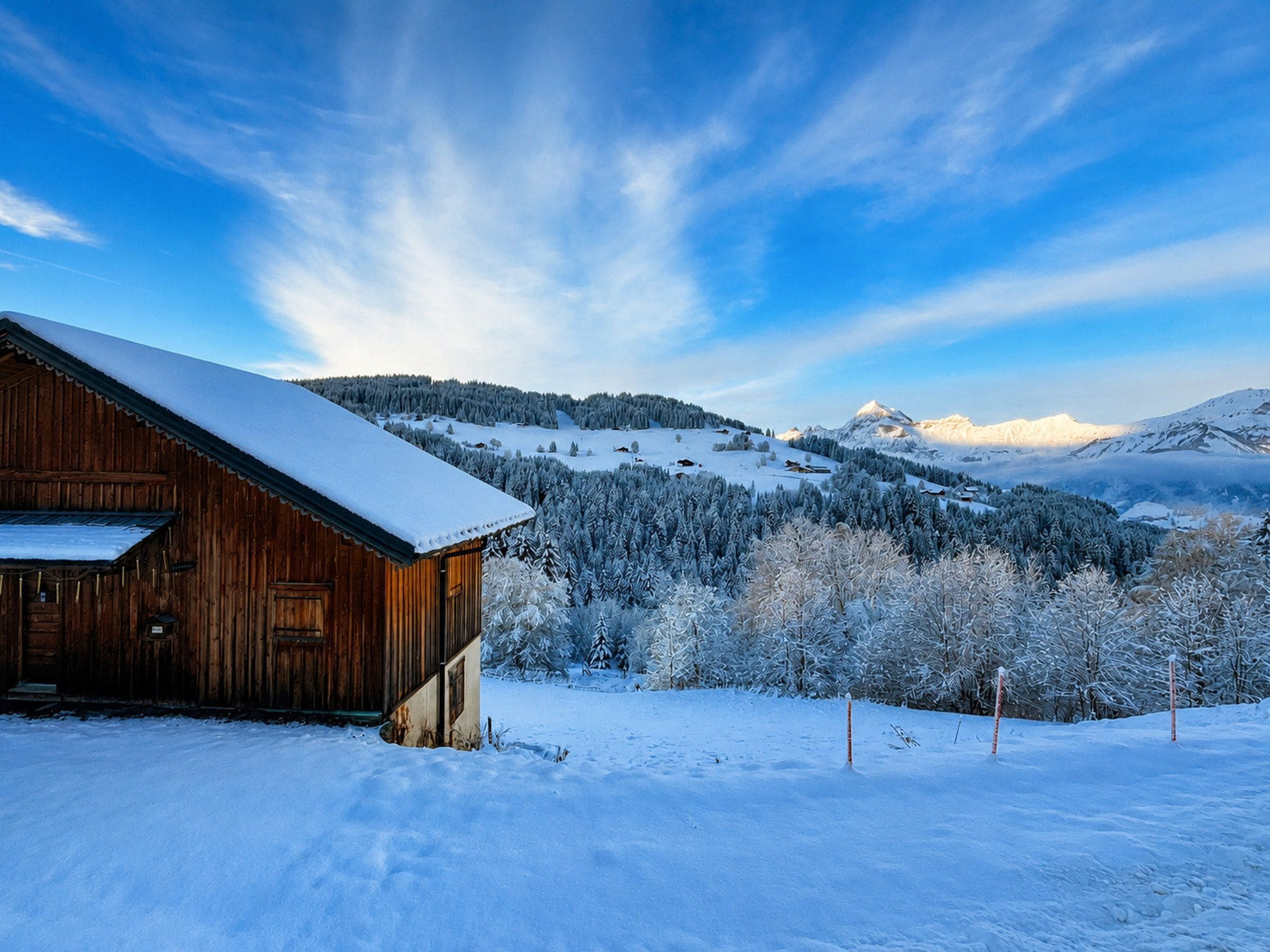 A wooden cabin with a snow-covered roof, surrounded by a snowy landscape and a mountain range.