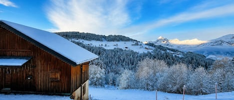 A wooden cabin with a snow-covered roof, surrounded by a snowy landscape and a mountain range.
