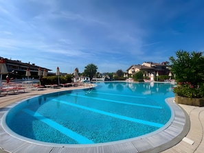 Una gran piscina cristal·lina amb gandules i ombrelles, envoltada d'edificis i vegetació.