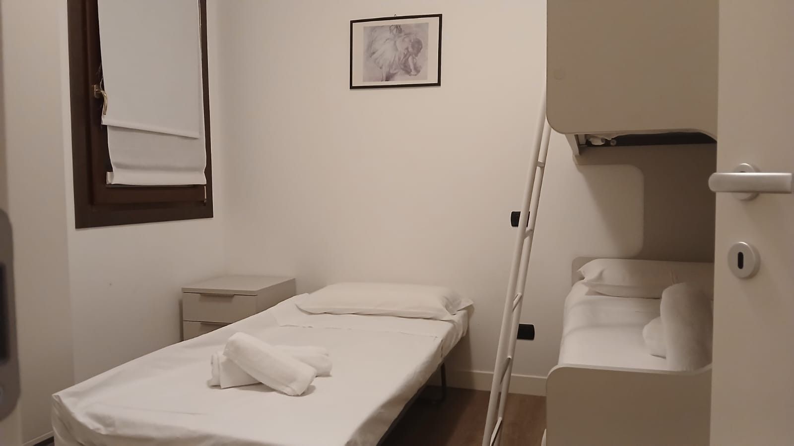 Ein Zimmer mit einem Einzelbett, einem Nachttisch, einer kleinen Kommode, einem gerahmten Bild an der Wand und einer Tür zu einem Badezimmer.