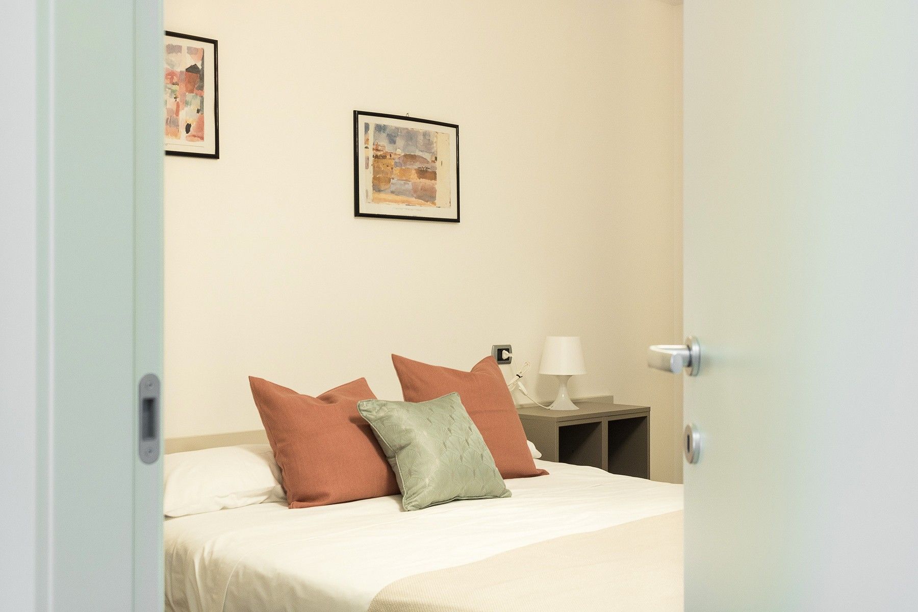 Habitación de hotel con cama, almohadas, una mesita de noche, lámpara y dos cuadros en la pared.