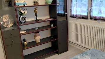 Ein Schlafzimmer mit Bett, einer Garderobe mit Ablagefächern, einem Heizkörper und einem Fenster mit Vorhängen.