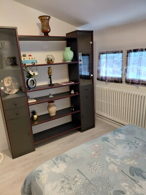 A bedroom with a bed, a wardrobe with shelves, a radiator, and a window with curtains.