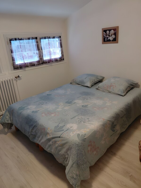 A bedroom with a bed, two pillows, a window with curtains, a radiator, and a wall-mounted picture.