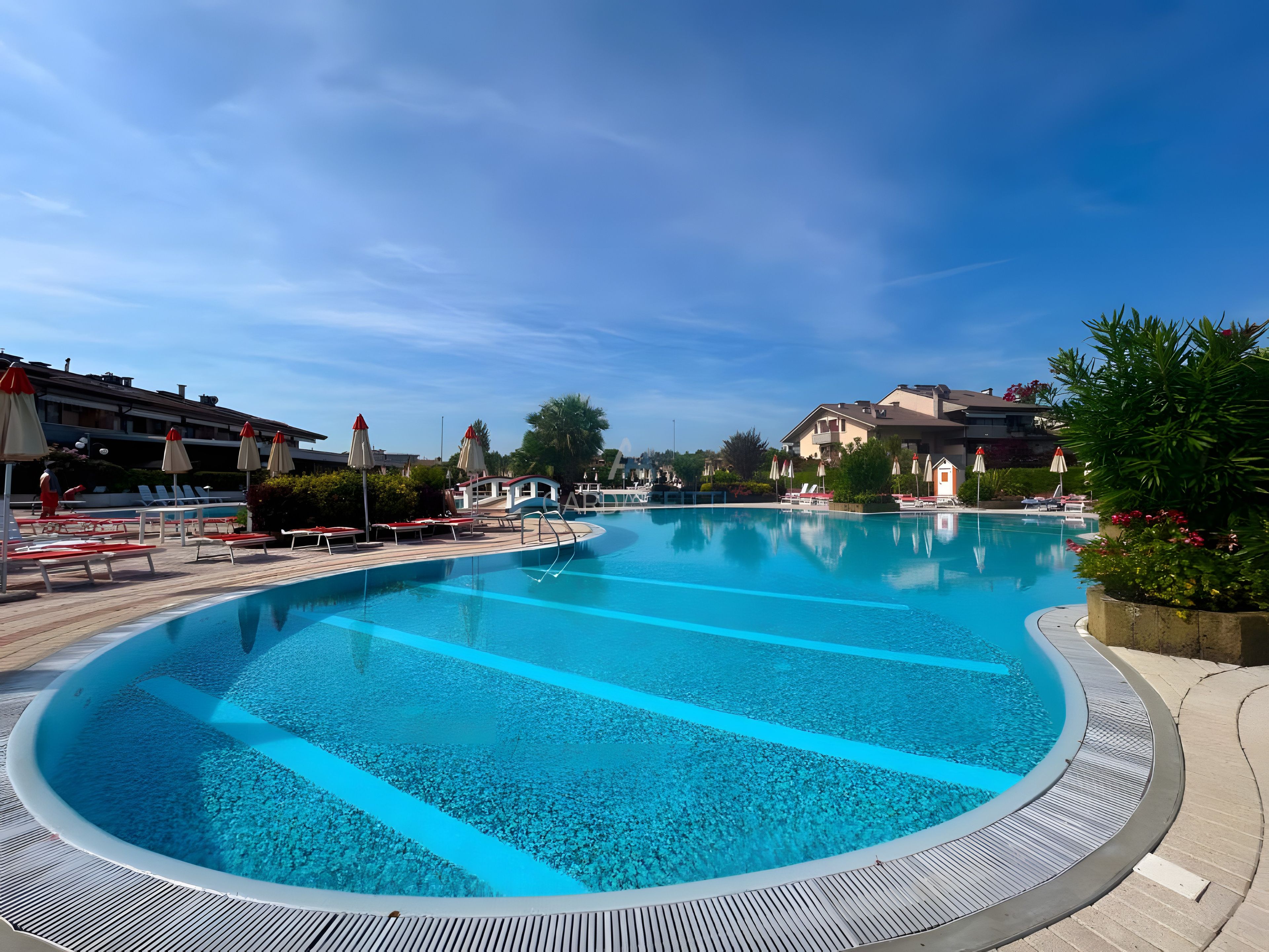 Ein großer, klares Schwimmbecken mit Liegestühlen und Sonnenschirmen, umgeben von Gebäuden und Grünflächen.
