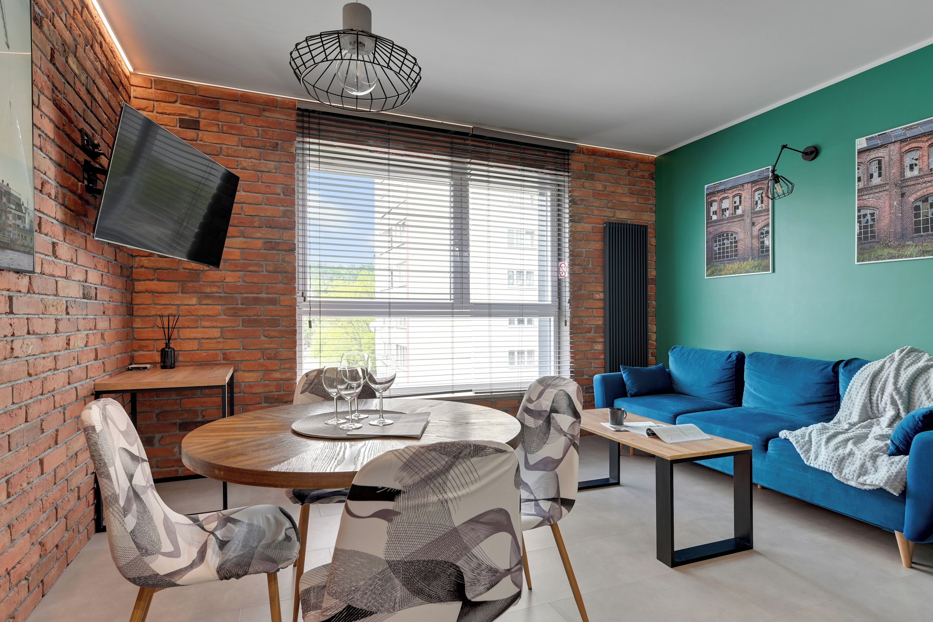 Een moderne woonkamer met een bakstenen muur, een ronde houten tafel, een blauwe bank en een wandgemonteerde televisie.