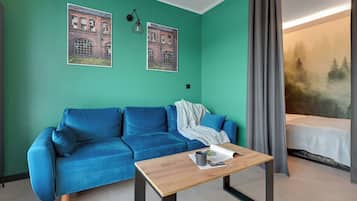A room with a teal wall, a blue sofa, a wooden coffee table, and a bed with a forest view.