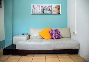 A room with a light-colored sofa against a teal wall, a framed picture above, and a small table with magazines. - Perfect 1-bedroom studio with AC in soothing San Juan (San Juan)