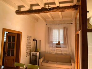 Una habitación con una viga de techo de madera, un sofá, un radiador y una ventana con cortinas traslúcidas.