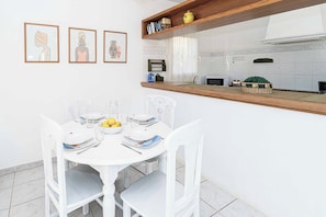 A dining area with a white table set for four, white chairs, and a kitchen in the background. - Villa Kiwi - Two Bedroom Villa, Sleeps 4 (El Tosalet)