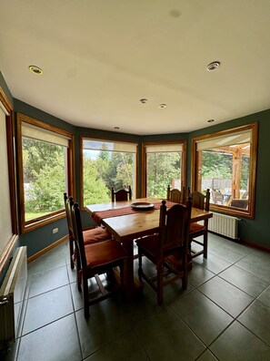 Ein Essbereich mit einem runden Tisch und Stühlen, großen Fenstern mit Blick auf Grünflächen und einem gefliesten Boden.