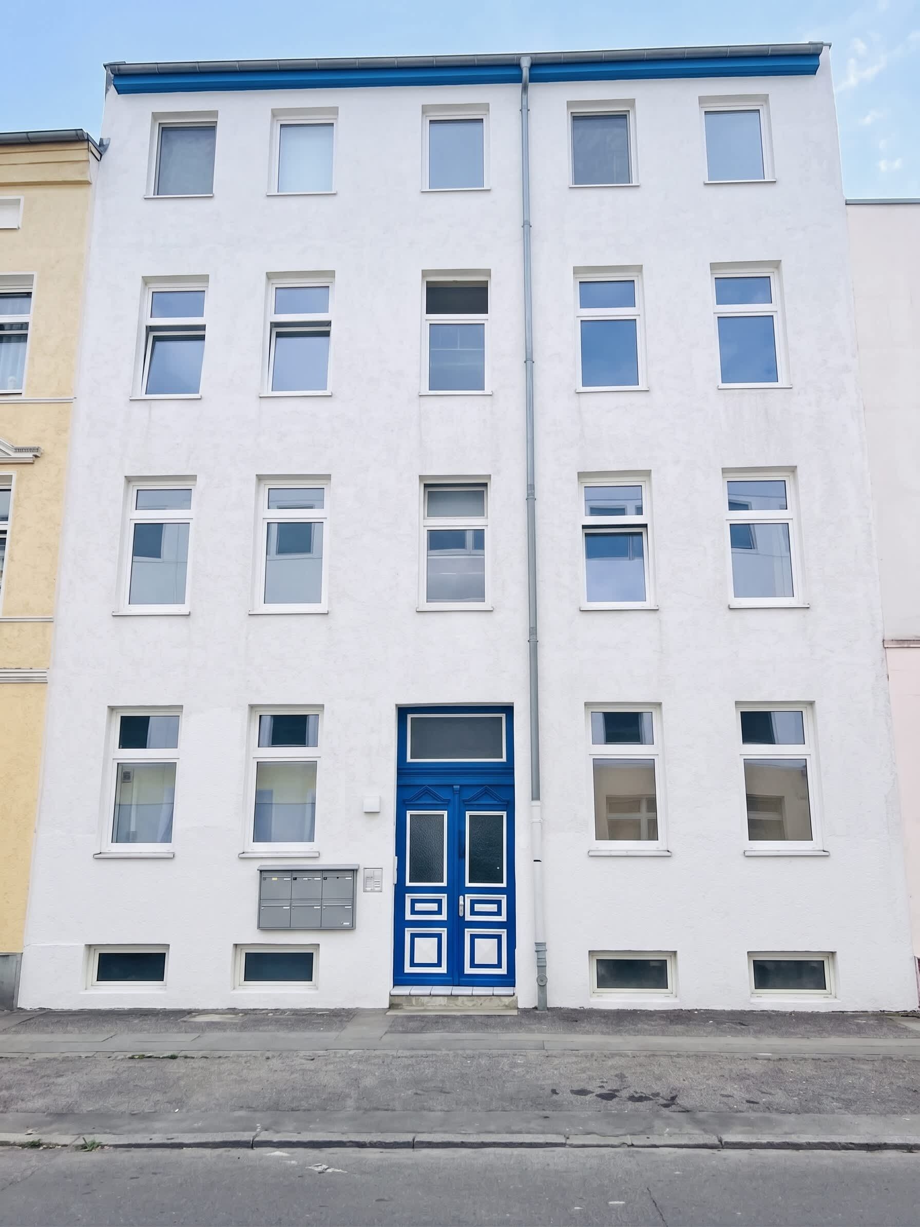 Ein mehrgeschossiges Gebäude mit einer blauen Eingangstür, zahlreichen Fenstern und einer sichtbaren Garagenöffnung.