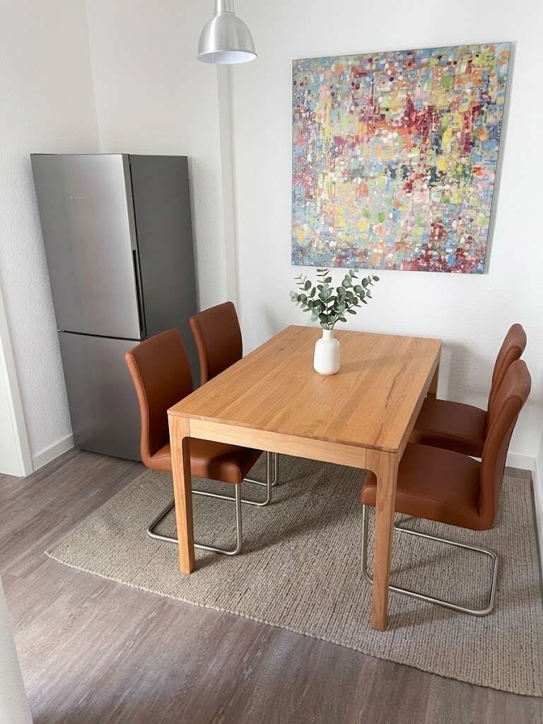 Ein Essbereich mit einem Holztisch und vier Stühlen, einer kleinen weißen Vase mit einer Pflanze sowie einem farbenfrohen, abstrakten Gemälde an der Wand.