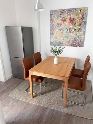 Et spiseområde med et træbord, fire stole, en lille hvid vase med en plante og et farverigt abstrakt billede på væggen.