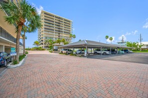 A hotel entrance with a covered parking area, a multi-story building in the background, and palm trees. - Beachfront Community w/ Pool, Workout Room + Tennis - Mansions By the Sea 406B (TREASURE ISLAND)