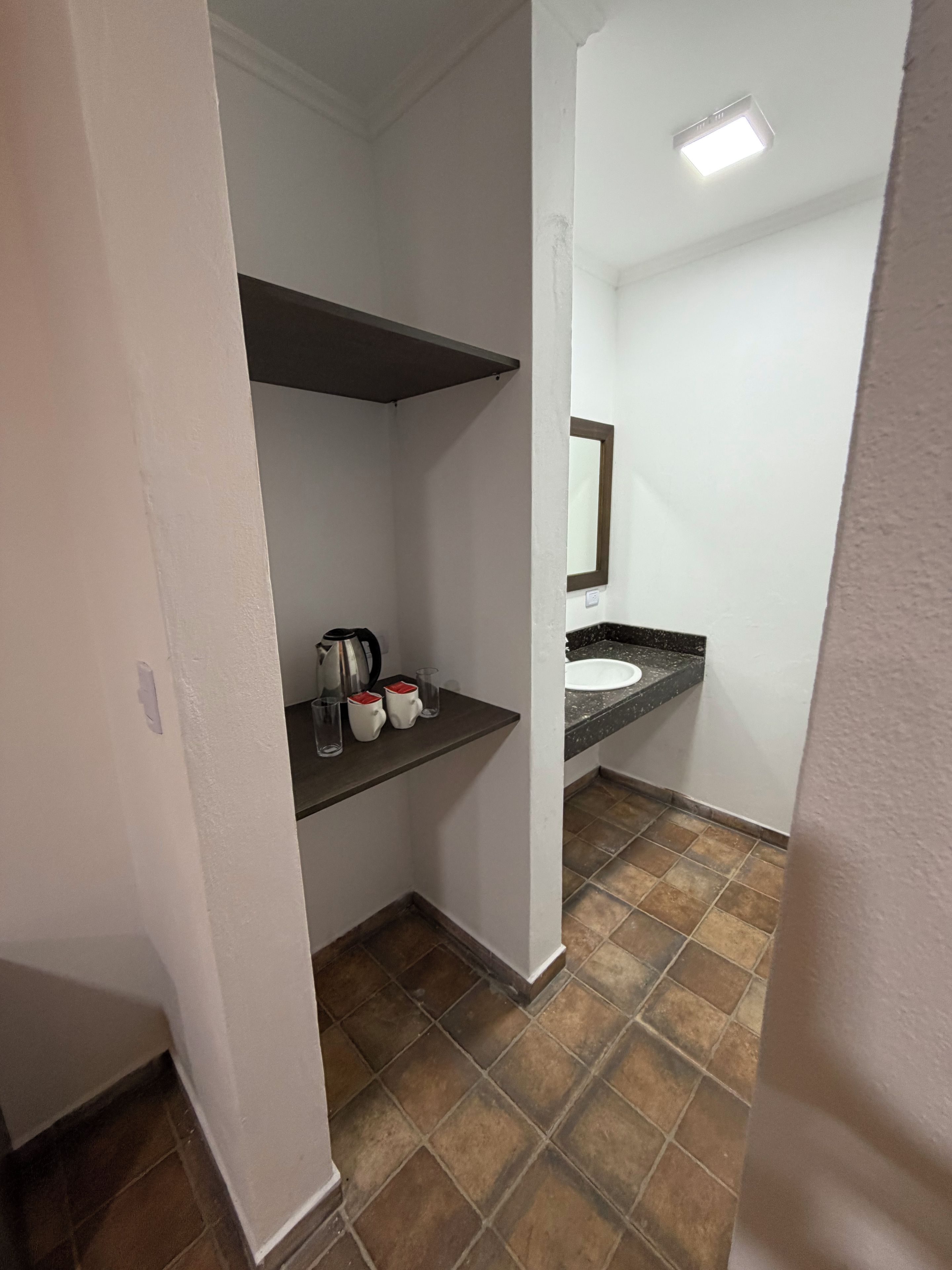 Casa de banho com lavatório branco, espelho, prateleira com chaleira e chávenas, e pavimento em mosaico castanho.