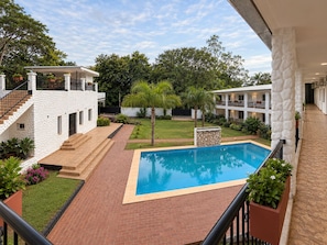 Uma área de piscina com piscina azul límpida, rodeada por um pátio de tijolos, e adjacente a um edifício branco com varanda.