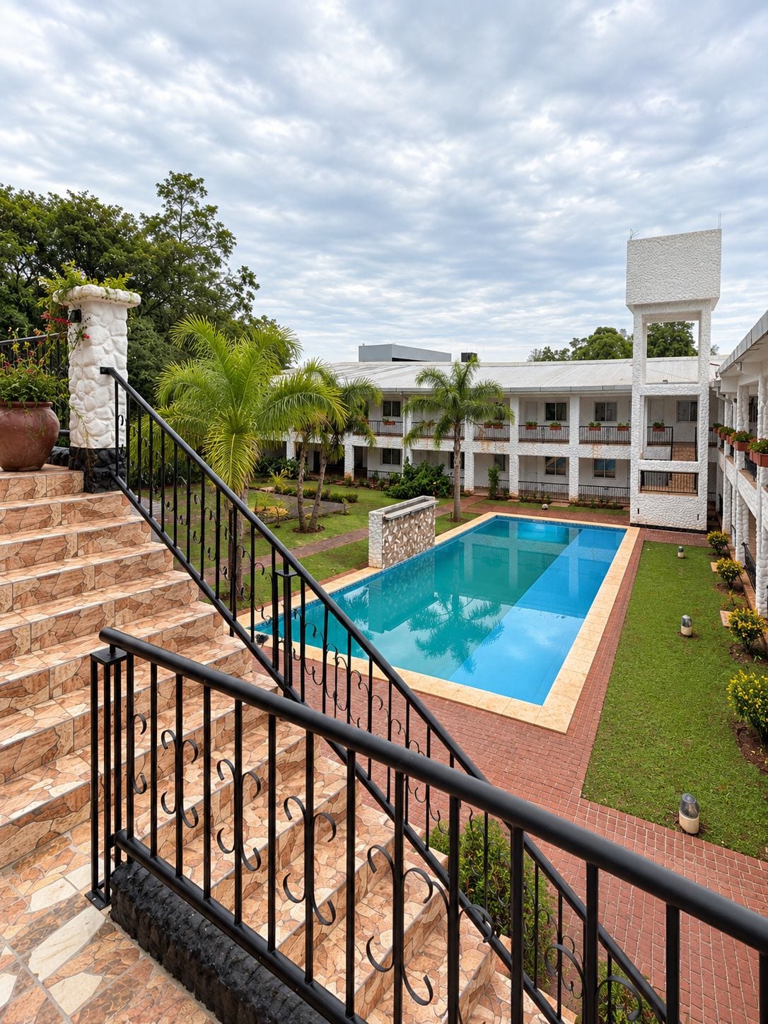 A hotel with a swimming pool, a staircase with a railing, and a garden area with palm trees.