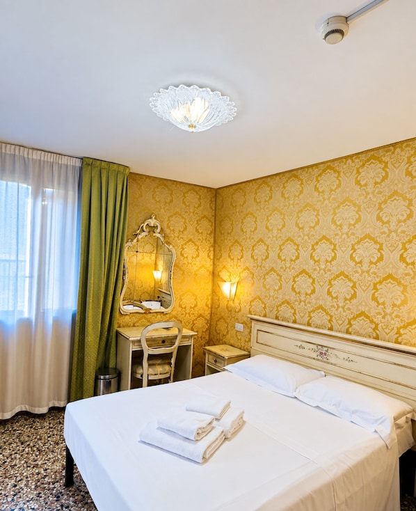 A hotel room with a bed, a desk, a chair, a mirror, a lamp, and a ceiling light fixture. - Double Room With Canal View (Venezia)