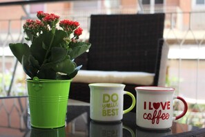 Ein grünes Kübelpflanzen mit roten Blüten, eine weiße Tasse mit grünem Text und eine rote Tasse mit weißem Text auf einem Glastisch.