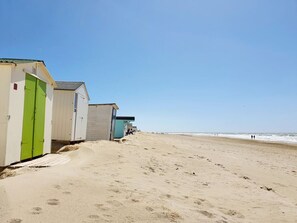 Ein Strand mit einer Reihe von Strandhütten, einige davon mit Türen, und einem wolkenlosen Himmel.