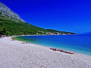 Ein Kiesstrand mit klarem, blauen Wasser, ein Berg im Hintergrund und einige Personen, die sich sonnen.