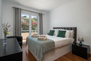 A hotel room with a bed, a desk, a chair, a nightstand, and a view of a balcony with flowers. - Apartment in Caniço near facilities|Boeiro Living (Caniço)