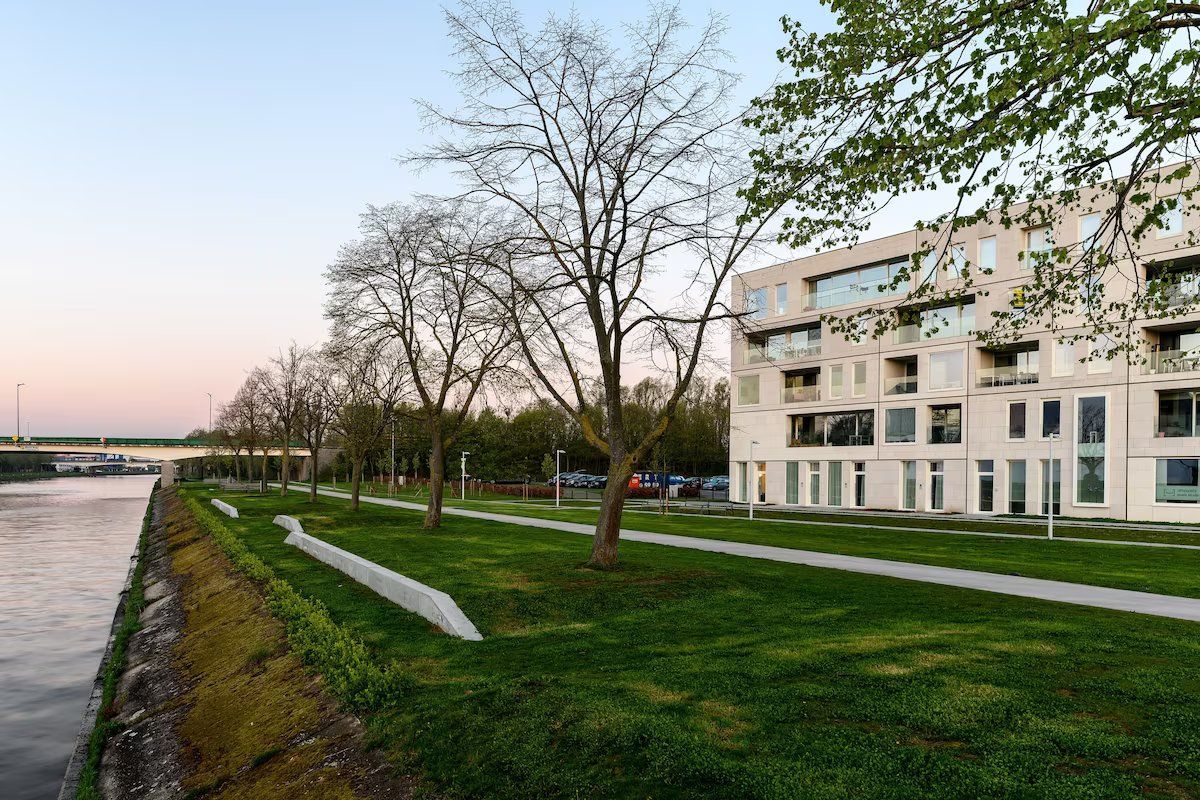 Een modern gebouw met een plat dak en grote ramen, gelegen naast een rivier met een verharde pad en bomen.