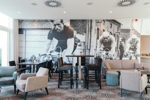A modern hotel lobby with a large mural of cyclists, comfortable seating, and a patterned carpet.