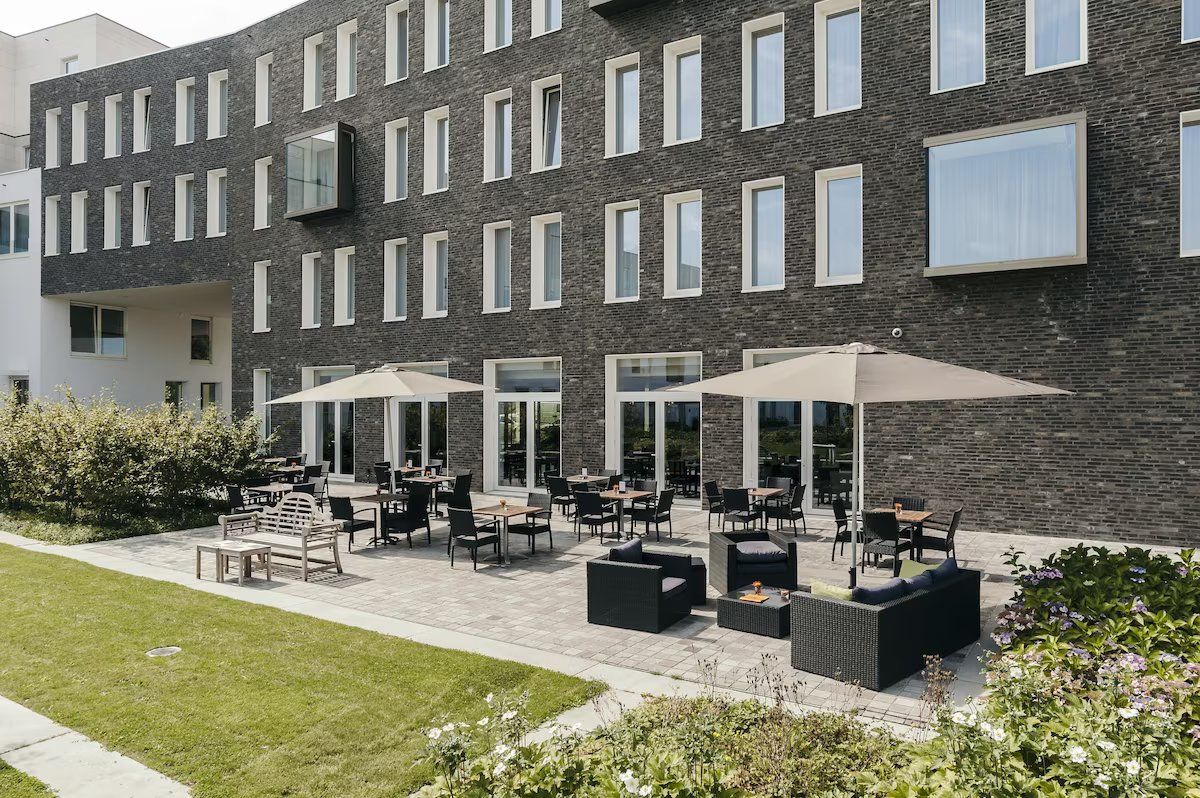 Un bâtiment moderne avec une façade en briques, un espace extérieur aménagé avec des tables et des parasols, et une pelouse bien entretenue.