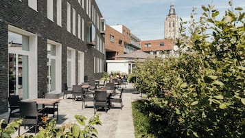 An outdoor patio area with tables and chairs, surrounded by greenery and a brick building.