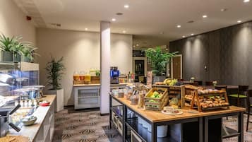 A hotel breakfast area with a counter displaying various food items, a coffee machine, and a seating area with tables and chairs.