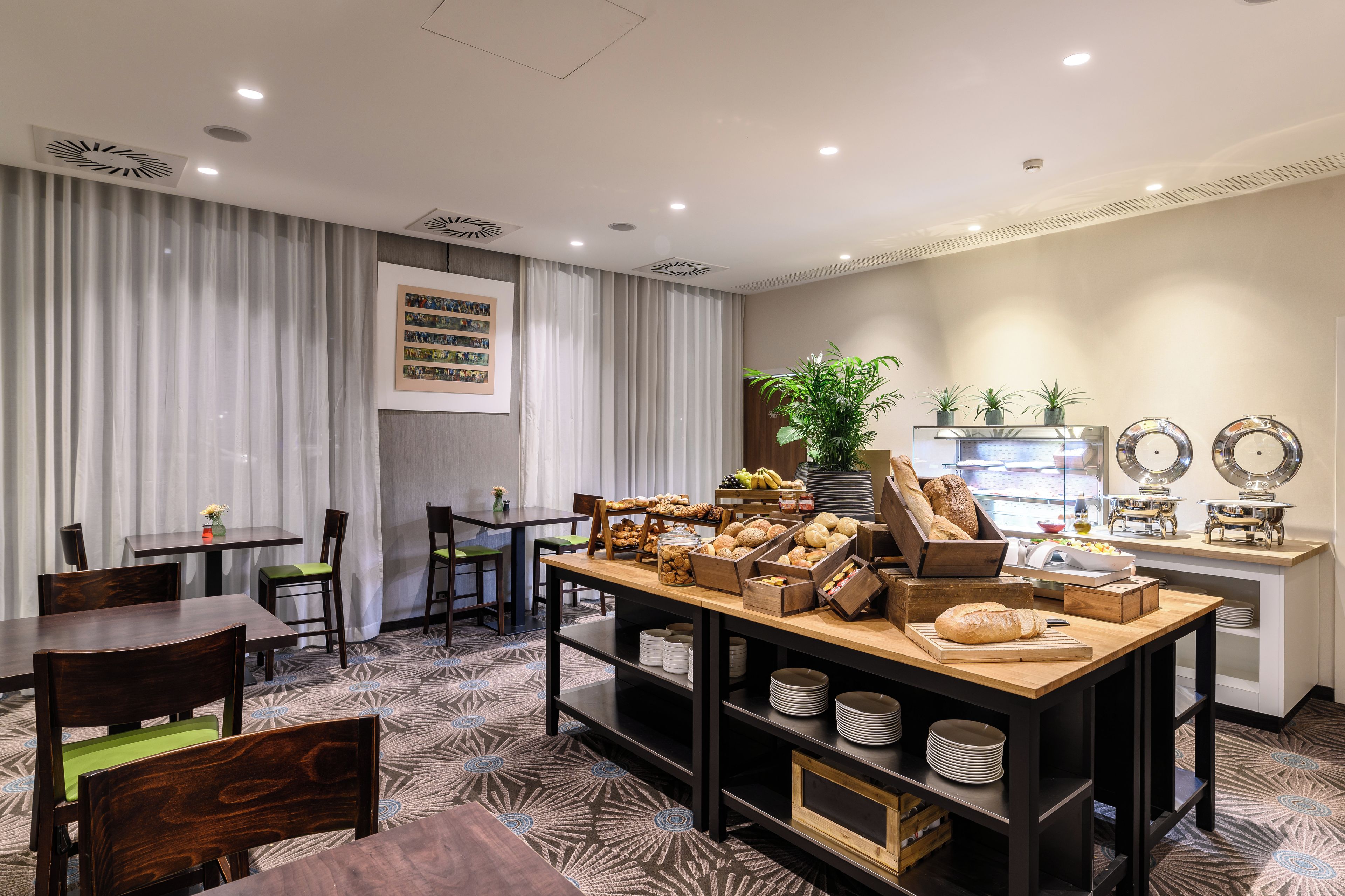 A dining area with a buffet setup, including bread, fruits, and various dishes. There are dining tables and chairs, a plant, and a wall with framed pictures.