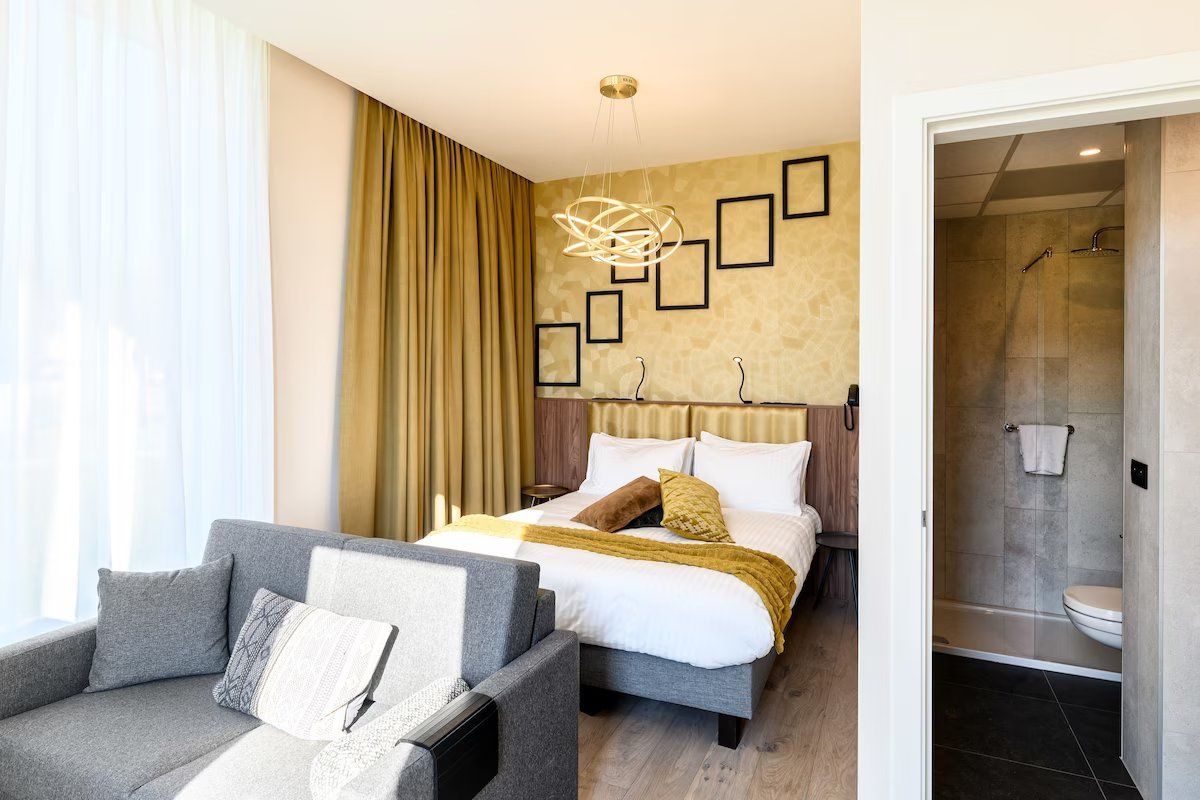 A modern hotel room with a bed, a sofa, a shower, and a view of the city.